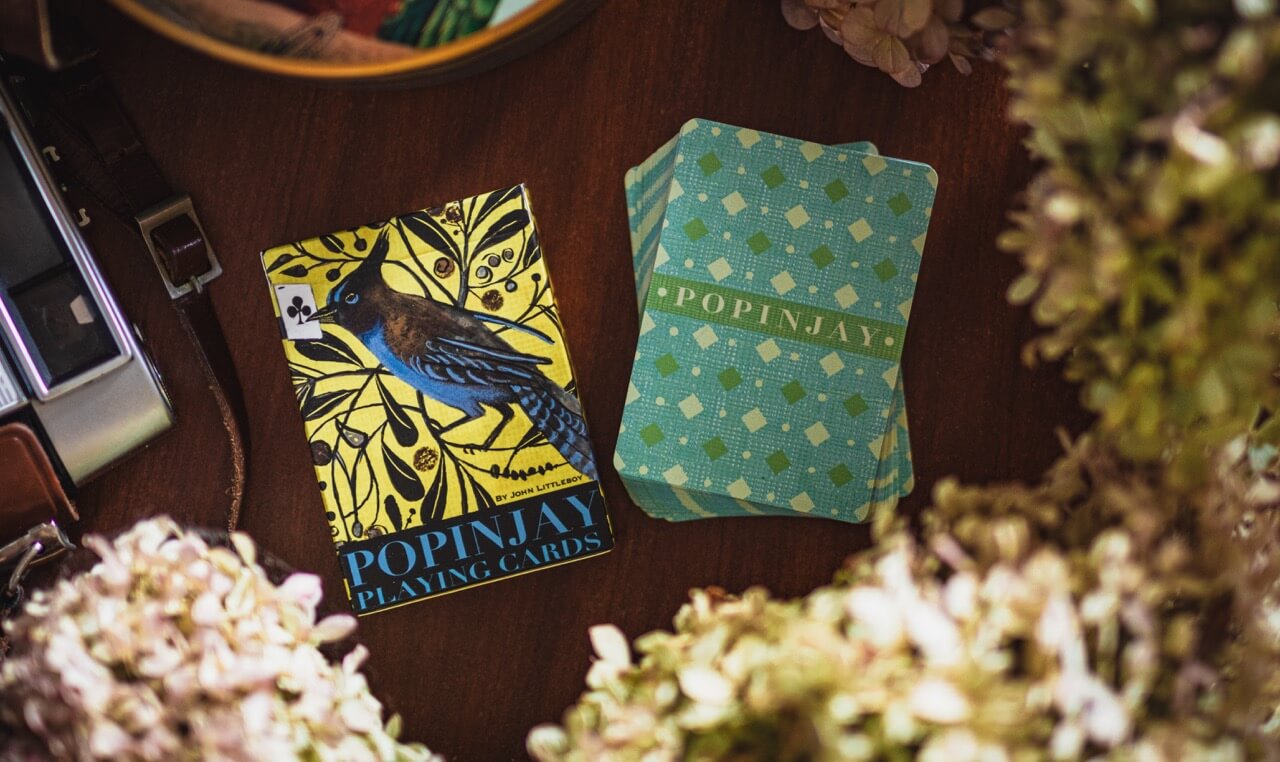 Two playing card decks from Popinjay on a wooden surface with flowers in the foreground.